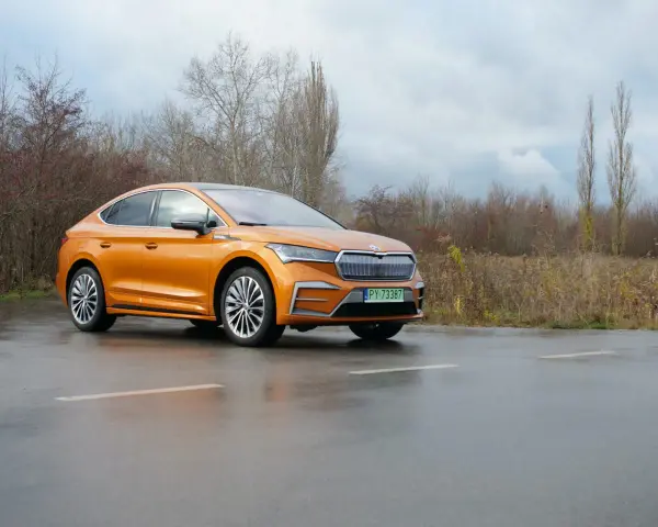 Pomarańczowa Skoda Enyaq Coupe na mokrej drodze, zarośla w tle.