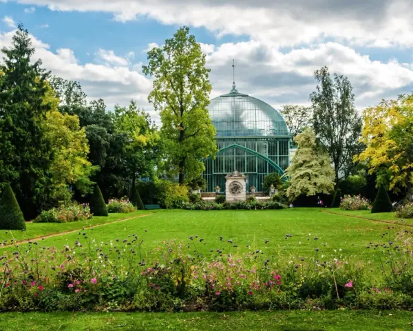 Un jardín exuberante en el Bois de Boulogne, con una gran estructura de cristal verde y un cielo azul con nubes.