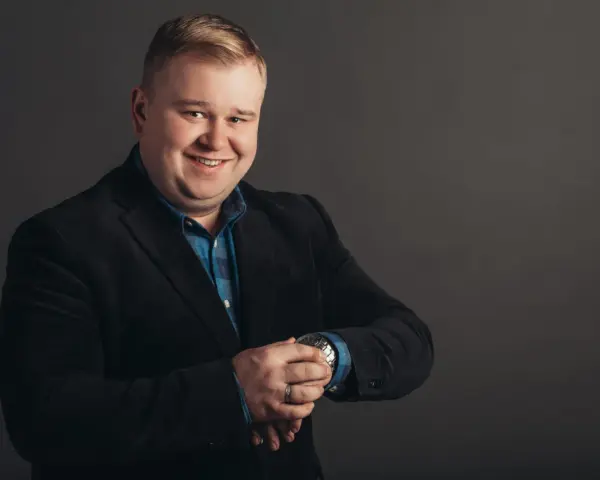 Uśmiechnięty mężczyzna w czarnym garniturze i niebieskiej koszuli, sprawdzający zegarek. Podpowiadamy, jak dobrać garnitur dla pana z brzuszkiem.