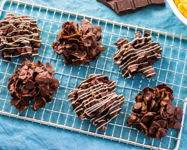 Sei dolcetti croccanti al cioccolato, simili a rose del deserto, raffreddano su una griglia. Alcuni sono decorati con sottili fili di cioccolato bianco.