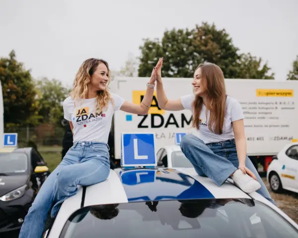 Dwie uśmiechnięte dziewczyny świętują sukces na dachu samochodu szkoleniowego. W tle ciężarówka z informacją o kursach na różne kategorie prawa jazdy.