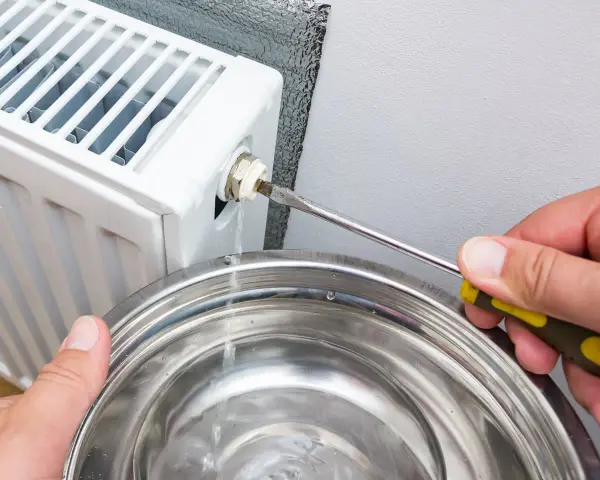 Osoba odkręca odpowietrznik grzejnika śrubokrętem, a woda kapie do metalowej miski. To pokazuje, jak odpowietrzyć grzejnik.