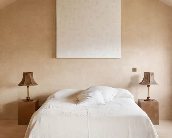 Camera da letto aesthetic con pareti color sabbia, letto bianco e due lampade in vimini. Un quadro astratto completa l'arredo.