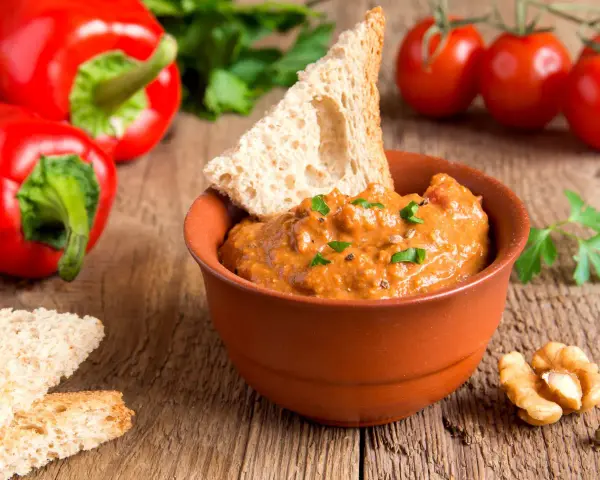 Un saporito pesto di peperoni in una ciotola di terracotta, accompagnato da crostini, noci e peperoni rossi freschi.
