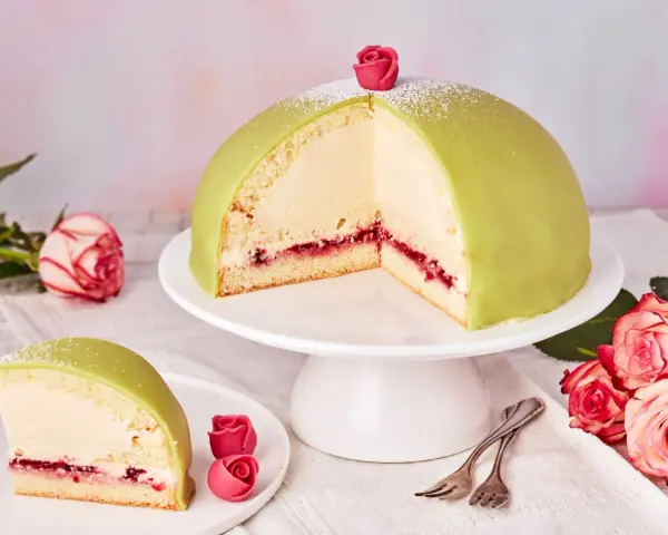 Ein Anschnitt eines grünen Prinzessinnenkuchens mit Sahnefüllung und Himbeermarmelade, verziert mit einer Zuckerrose. Ein Stück liegt auf einem Teller daneben.