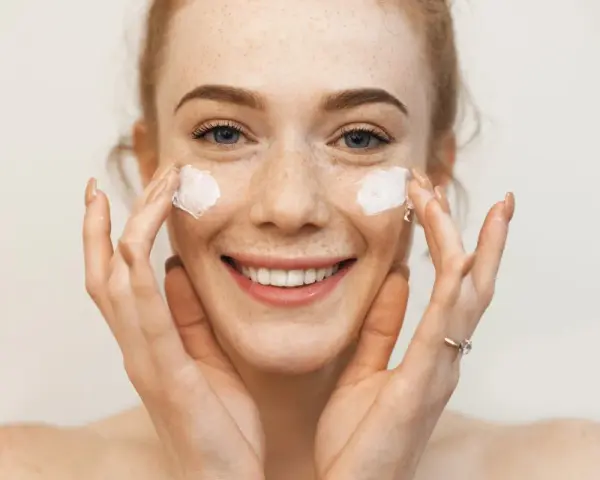 Uśmiechnięta dziewczyna z piegami nakłada krem na twarz. Sudocrem na co? Na suchą skórę, podrażnienia, zaczerwienienia.