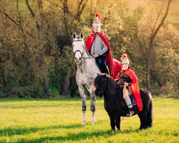 Zwei Reiter in römischer Kleidung, ein Erwachsener auf einem Schimmel und ein Kind auf einem Pony, reiten auf einer sonnigen Wiese. Ein fröhlicher Anblick, wie bei einem Martin Fest.