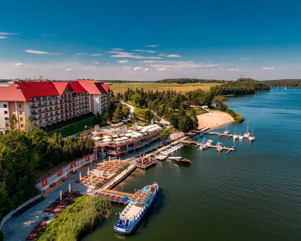 Hotel na wyspie Mikołajki z czerwonym dachem, otoczony zielenią, z mariną pełną łodzi i statkiem wycieczkowym na jeziorze.