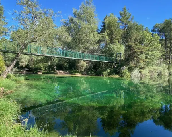 Puente verde cruza un río de aguas cristalinas y verdes, reflejando el bosque exuberante. Un lugar idílico para explorar el **alto tajo**.