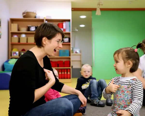 Eine Frau lacht und gestikuliert, während sie mit einem Kleinkind sprechen übt. Ein Junge und ein Mann sitzen im Hintergrund.