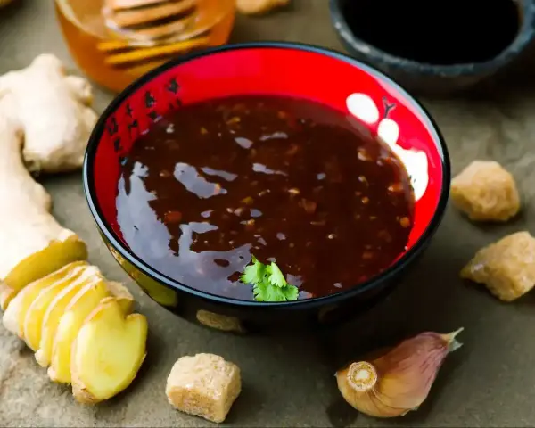 Miseczka z ciemnym sosem teriyaki, obok imbir, czosnek, cukier trzcinowy i miód.