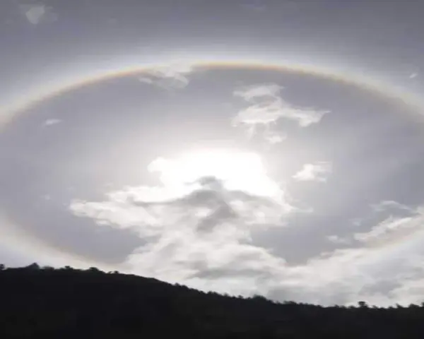 Un alone arcobaleno circonda il sole tra le nuvole, creando un effetto magico sopra una collina scura.