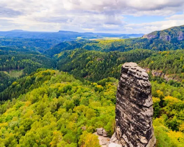 Skalny filar góruje nad zielonymi lasami w Czechach, blisko granicy. Krajobraz pełen jest drzew i skalistych zboczy.