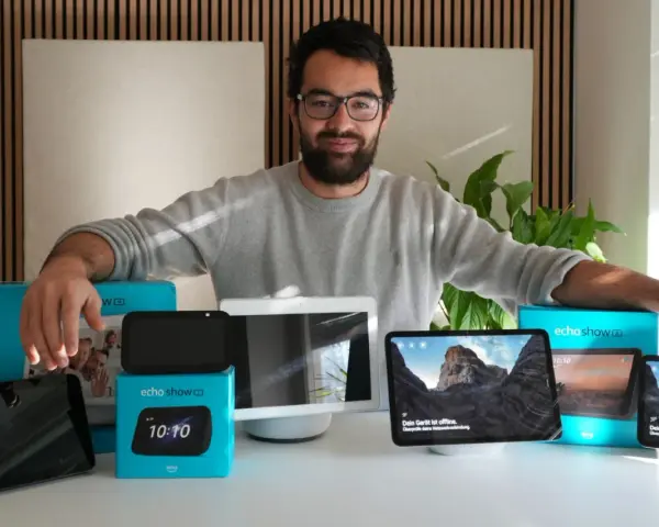 Mann präsentiert verschiedene Echo Show Generationen. Mehrere Geräte und Verpackungen sind auf dem Tisch zu sehen.