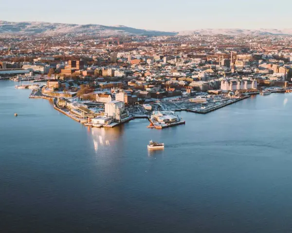 Widok na zimowe Oslo z wody. Mała łódka płynie po spokojnym akwenie, w tle rozciąga się miasto i ośnieżone wzgórza.