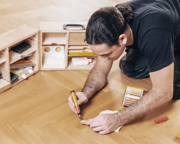 Mann repariert Parkett mit einem Reparaturstift und einer Farbauswahl. Werkzeugkasten mit Zubehör daneben.