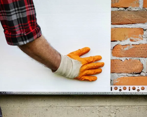 Ręka w pomarańczowej rękawicy przykłada płytę pir do ceglanej ściany.
