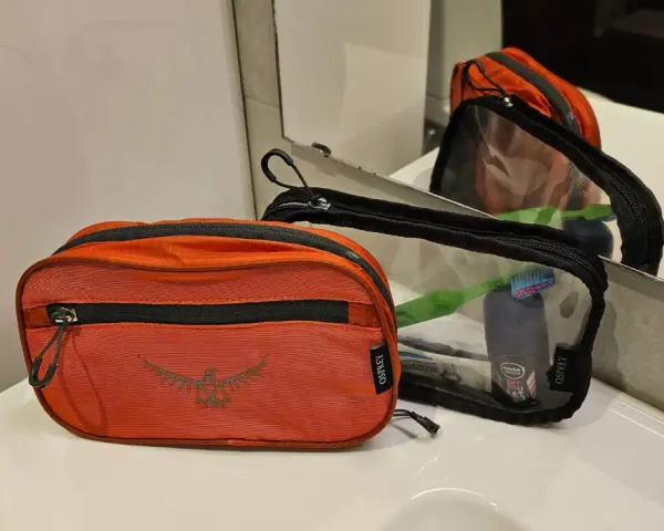 Två Osprey-necessärer, en orange och en genomskinlig, ligger på en vit bänkskiva. Den genomskinliga necessären innehåller tandborste och deodorant.