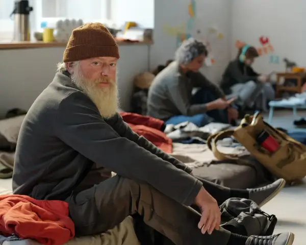 Älterer Mann mit Bart und Mütze sitzt auf dem Boden, umgeben von anderen Menschen. Eine Szene, die die Frage aufwirft: Wohin ohne Geld und Wohnung?