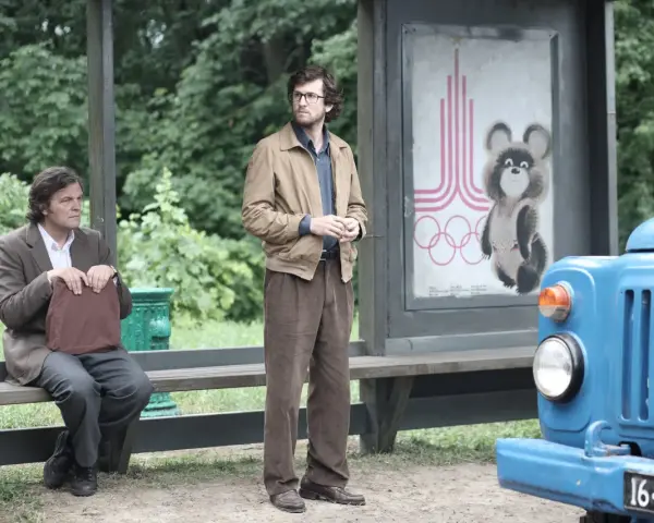 Deux hommes attendent à un arrêt de bus, un panneau des JO de Moscou 1980 avec Misha l'ours en arrière-plan. L'affaire Farewell se déroule.
