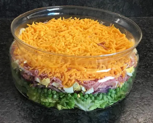 Une salade 7 étages colorée dans un grand bol en verre, avec des couches de laitue, petits pois, oignons rouges, jambon, œufs durs et fromage râpé.