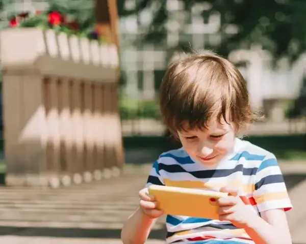 Chłopiec w kolorowym T-shircie ogląda film dla małych dzieci na żółtym telefonie, siedząc na zewnątrz.