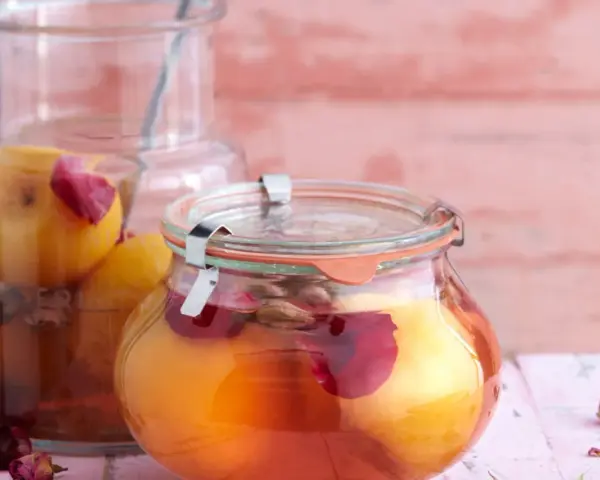 Zwei Gläser mit eingelegten Pfirsichen, verziert mit Rosenblättern und Gewürzen, auf einem rosa Holztisch.