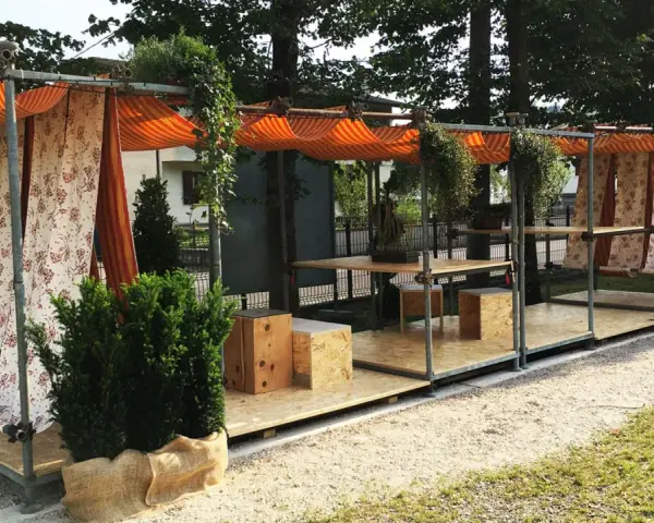 Struttura esterna con tende a righe e floreali, tavoli e sedute in legno. Un esempio di come costruire un gazebo con tubi innocenti, pratico e decorativo.