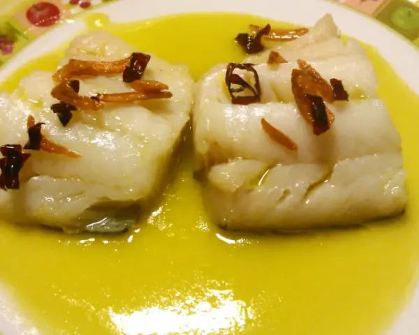 Dos trozos de bacalao al pil pil, cubiertos con ajo frito y guindilla, nadan en una salsa dorada sobre un plato decorado.