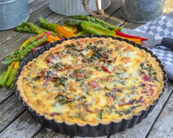 Herzhaftes Mangold Quiche Rezept, frisch gebacken und bereit zum Genießen. Bunte Mangoldstiele liegen daneben.