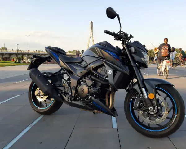 Czarny motocykl Suzuki GSX-S 750 z niebieskimi akcentami na felgach i owiewkach, zaparkowany na placu z widocznym w tle mostem i ludźmi.