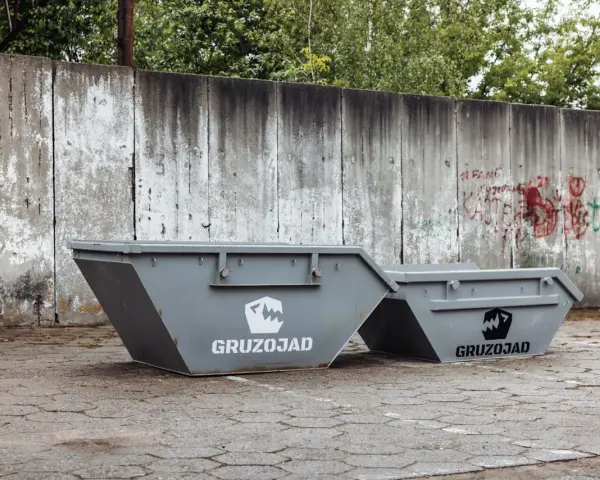Dwa szare kontenery na śmieci zmieszane, z logo GRUZOJAD, stoją przed zniszczonym betonowym murem pokrytym graffiti.