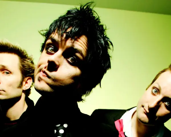 Trzej członkowie Green Day: Billie Joe Armstrong, Mike Dirnt i Tré Cool, pozują do zdjęcia na zielonym tle.