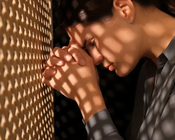 A woman in a confessional booth, hands clasped, seeking guidance on how to ask God for forgiveness.