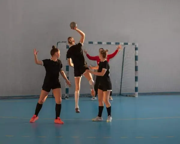 Piłkarki ręczne w akcji. Jedna z nich skacze do rzutu, podczas gdy koleżanki z drużyny i przeciwniczka obserwują. To dynamiczna gra pasywna w piłce ręcznej.