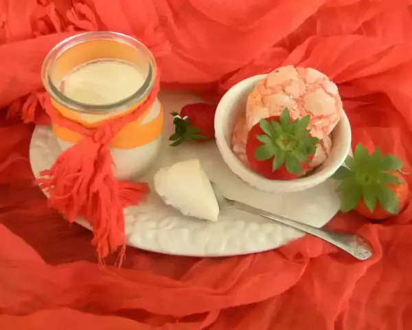 Yogur cremoso en frasco con fresas y trozos de yogur en un plato blanco sobre tela naranja.