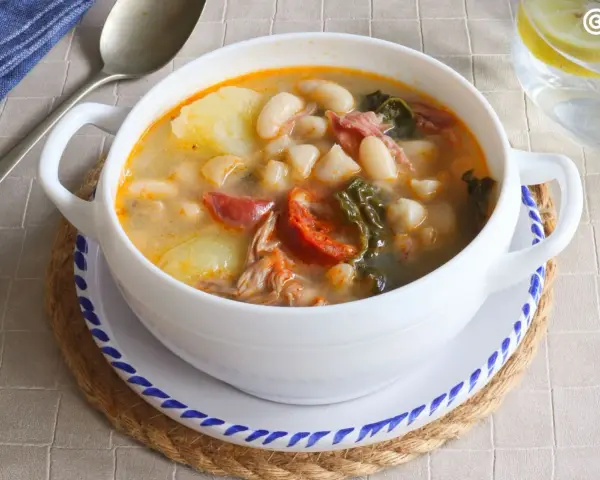 Un tazón blanco de caldo gallego con patatas, alubias, chorizo y grelos.