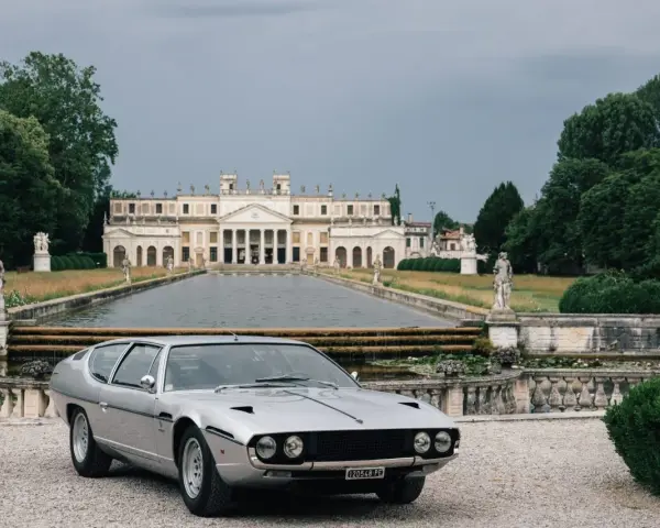 Srebrny Lamborghini Espada z lat 70. przed okazałą willą z basenem i rzeźbami.