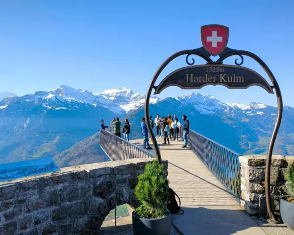 Tourists enjoy the breathtaking panorama from the Harder Kulm viewpoint, with snow-capped mountains under a clear blue sky.