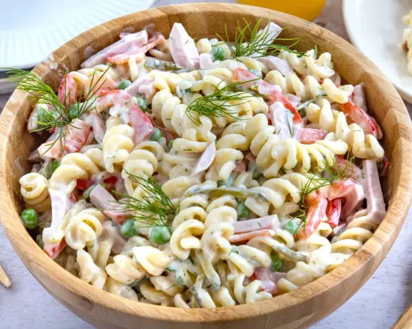 Ein großer Holzteller voller cremigen Nudelsalat mit Schinken, Erbsen und Paprika. Mit Dill garniert, sieht er aus wie der beste Nudelsalat!