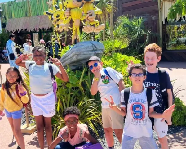 Des enfants joyeux font des signes de paix devant une attraction, un avis "supernova juniors" est visible.