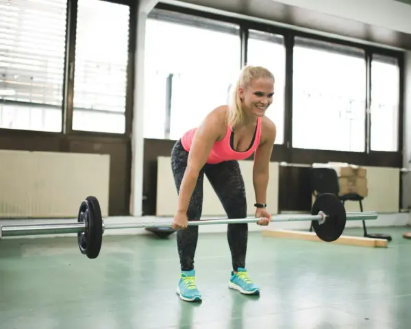 En kvinna lyfter skivstång i ett gym. Hon ler och ser fokuserad ut, en bra start på hur blir man stark.