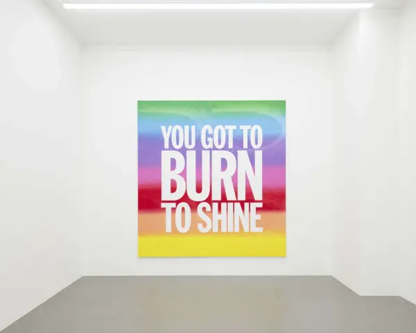 Œuvre de John Giorno dans une galerie, avec le message "YOU GOT TO BURN TO SHINE" sur un fond arc-en-ciel.
