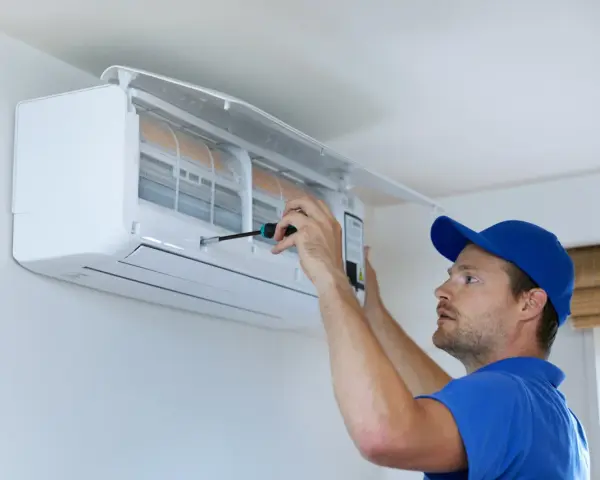 Un technicien en bleu utilise un tournevis pour ajuster un climatiseur mural, assurant le bon fonctionnement pour recharger clim maison.