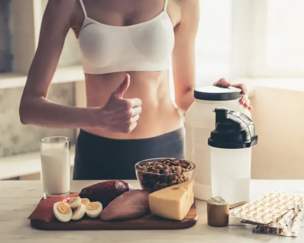 Kobieta z kciukiem w górę prezentuje produkty wspierające dietę wysokobiałkową: mięso, rybę, jajka, ser, orzechy, mleko i suplementy.