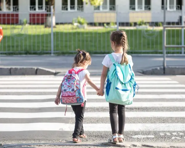 Dwie dziewczynki z plecakami idą za rękę przez pasy. Od ilu lat dziecko może samo chodzić po mieście?