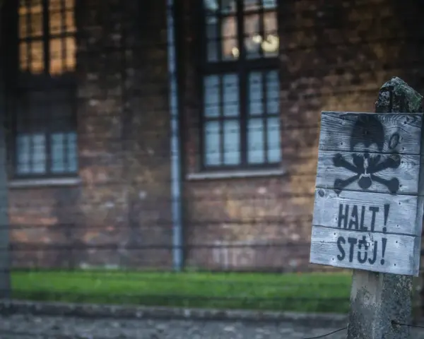 Tablica z symbolem czaszki i piszczeli oraz napisem "HALT! STÓJ!" na tle ceglanego budynku i drutu kolczastego, przypominająca o grozie obozów koncentracyjnych.