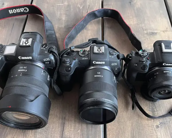 Trzy aparaty Canon EOS R, RP i R5 leżą obok siebie na drewnianym blacie. To **pełna klatka** sprzętu fotograficznego.
