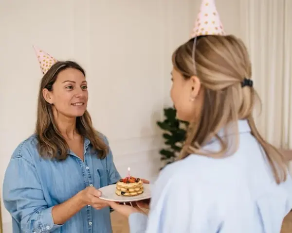 Dwie kobiety w czapkach urodzinowych. Jedna podaje drugiej mały tort ze świeczką. To miły gest, jak potwierdzić przybycie na urodziny.