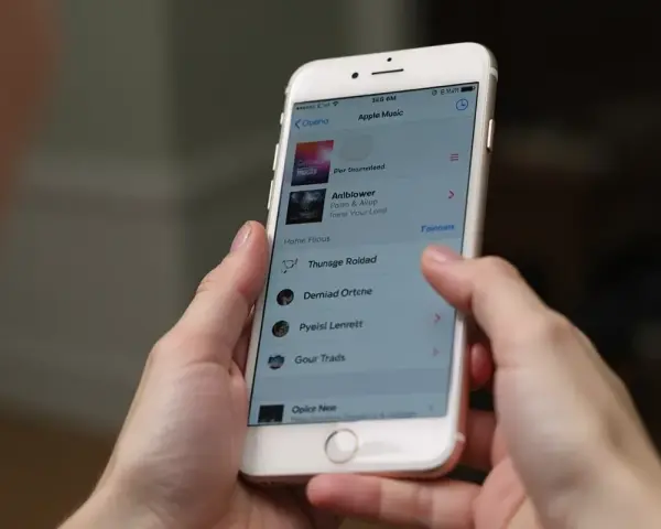 Ręce trzymają iPhone'a z otwartą aplikacją Apple Music. Widoczna lista utworów, która może pomóc w nauce, jak pobrać muzykę na iPhone'a.